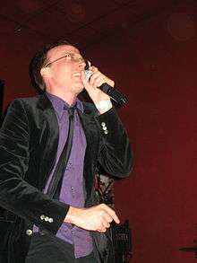 Photograph of a man in a purple shirt and black suit holding a microphone.