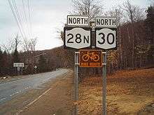 Signage for Route 28N and Route 30 heading northbound through a wooded community with a brown and white bicycle route sign