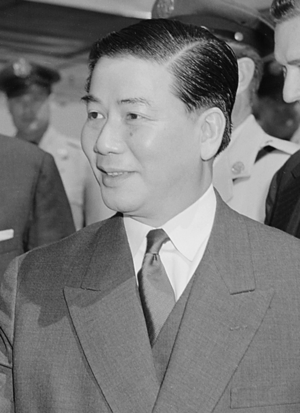 A portrait of a middle-aged man, looking to the left in a half-portrait/profile. He has chubby cheeks, parts his hair to the side and wears a suit and tie.