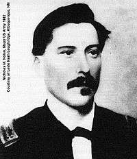 Black and white picture of a white male with a full dark mustache and squared goatee facing the camera and looking slightly to the right. He has dark swarthy hair neatly combed over a broad forehead. His dress uniform has a white collar with the front ends turned down with a small bow tie.