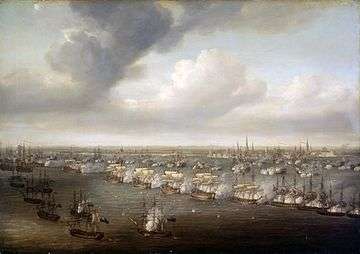 Bird's eye view of a naval battle, with sailing ships arranged in two parallel lines running diagonally from the top left to the bottom right of the painting. A small cluster of ships in the bottom left. A dark sea and a cloudy sky, the buildings and spires of a city visible in the background.