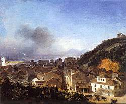 A painting of a view from a hilltop with a group of friars in the foreground, the buildings of a town between the foot of the hill and an adjacent hill, and sailing ships riding at anchor in the distance
