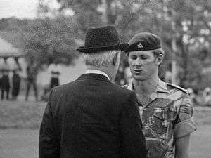 A black-and-white photograph of a soldier and a gentleman in a dark suit and hat shaking hands. The man in the suit is facing away from the camera. The soldier wears a dark-coloured beret with insignia on it and a camouflage shirt on which the sleeves are rolled up. A cross-shaped medal is prominent on his chest pocket. In the background, out of focus, four dark figures can be seen.