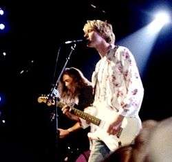 A side-view photograph of a blonde-haired man playing a guitar and singing into a microphone, along with a brown-haired man playing a bass guitar.