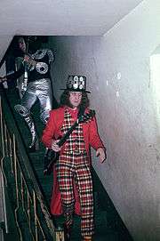 A man walks down a flight of stairs. He holds a guitar and wears a black hat studded with round metal plates. His vest and pants are chequered. His coat and shirt are plain red. His socks are striped.