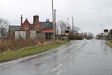 On a wet day, with rain falling, we are standing around 400 yards from a level crossing on a wide but not busy road.  The crossing, protected by lifting barriers, is next to the former station, a building in red brick with stone Mullions.