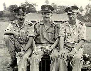 Three men in light-coloured uniforms with peaked caps seated on a jeep bonnet