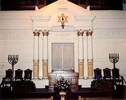 A white wall with an inset Torah ark at its center faces the viewer. Atop the ark is gold-colored Hebrew writing, and atop that a hanging lamp suspended from the center of a gold-colored Star of David. On each side of the ark are tall white columns, and beside them three ornate dark chairs and large seven branched menorahs. In front of the ark is a wooden pulpit.
