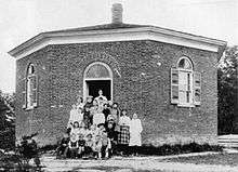 A black-and-white photo of students and their teacher in front of the Eight Square Schoolhouse, an eight-sided one-room schoolhouse, in Dryden, New York, in 1890
