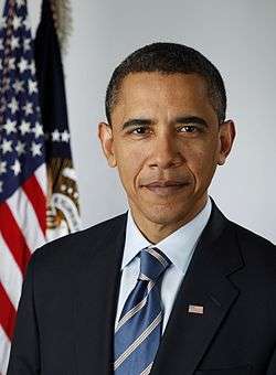 A man wearing a black suit and blue-striped tie, with the American flag in the background