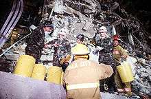 Several Air Force members and firefighters are clearing debris from the damaged building. Several yellow buckets are visible, which are being used to hold the debris. The destruction of the bombing is visible behind the rescuers.
