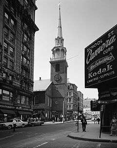On a city street, an old brick church with a tall steeple is surrounded by modern buildings.