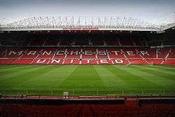 A stand of a football stadium. The seats are red, and the words "Manchester United" are written in white seats. The roof of the stand is supported by a cantilever structure. On the lip of the roof, it reads "Old Trafford Manchester".