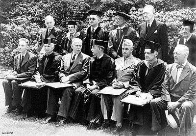 Thirteen men pose for a formal picture. Seven are sitting and six standing. There are all wearing either suits or academic gowns, except Bradley, who is in his Army uniform with a garrison cap.