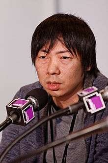 A photo of a middle aged Japanese man who is surrounded by microphones.