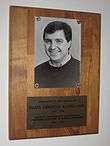 Brown wooden plaque hanged on a wall, bearing an inscription and a black and white photo of man in his forties.