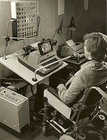  a black and white photograph showing a user sitting in front of a modified typewrite, there is grid of letters above the typewriter that appear to be lighting up in sequence, a tube goes from the users mouth to the machine