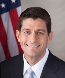 A portrait shot of Paul Ryan, looking straight ahead. He has short brown hair, and is wearing a dark navy blazer with a red and blue striped tie over a light blue collared shirt. In the background is the American flag.