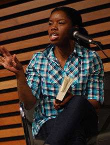 A woman is sitting in a chair and holding a book. She is speaking into a microphone to an unseen audience.