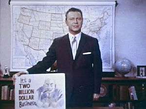 A still shot from the 1965 film Perversion for Profit showing a man in a suit holding a placard. The placard features a cartoonish image of a leering man and the phrase "It's a two billion dollar business"