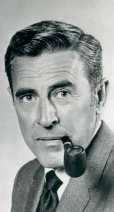 A man with dark hair, wearing a suit, including a tie also having a pipe in his mouth.