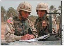 two men wearing three color desert combat uniforms, both with general insignia, look at a map and notebook atop a hood of a HMMWV.
