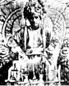 black and white photograph of a young cleanshaven man wearing an elaborate headdress and sitting on a throne. He is wearing robes and in front of him are some accoutrements on a table.