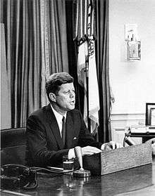 A black and white photograph of President John F. Kennedy speaking into a microphone at his desk in the Oval Office of the White House