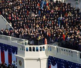 View of a large portion of a large ceremony with visible red, white and blue ornamentation and a crowd of attendees