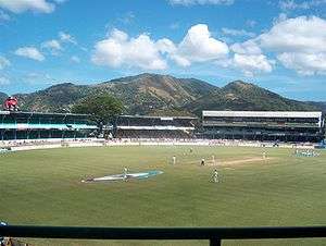  A stadium with people playing cricket. Large mountains are in the background, behind the stands