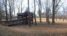 An open wooden structure with a ramp leading up to it is one the edge of a large open field, with large leafless trees around.