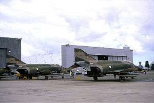 Two military jet fighters painted in camouflage livery parked in front of a large white building