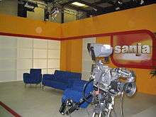 Orange, red and white TV studio, with blue chairs and gray camera