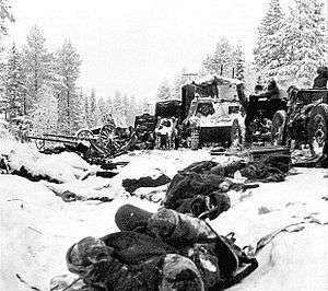 A road in the middle of forest and snow, a frozen body in front and destroyed vehicles in behind.