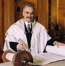 A gray-haired man with a full mustache faces the viewer, wearing a large skullcap, round wire-rimmed glasses, black suit with white shirt and patterned tie, and a full white prayer shawl with light gray stripes. On a table before him is an open Torah scroll; his left hand rests on one side of the scroll, and his right hand holds a silver pointer pointed at the words written on the scroll. A wall and part of a closed Torah ark are visible behind him.