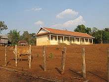 Small white building standing in a field of red earth. A cow wanders in the foreground.