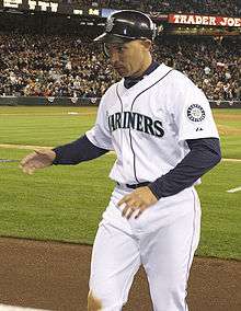 An olive-skinned man wearing a white baseball jersey with "Mariners" across the chest and a dark baseball cap