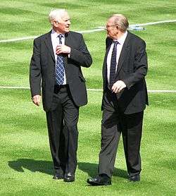 Two elderly gentlemen in suits, walking on a football pitch