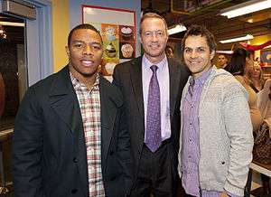 Three men stand together and smile. The man on the left is African American and wearing a plaid shirt with a black jacket. The man in the center is Caucasian and wearing a black suit with a light purple shirt and a dark purple tie, while the man on the right is Caucasian and wearing a purple checkered shirt with a grey sweater. A poster of cupcakes is visible on the wall behind them. People are talking to each other in the background.