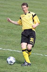 An in-action photograph of a man wearing a yellow football shirt, black shorts and yellow socks. The man has just passed the football to someone out of picture.