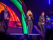 Madonna onstage wearing  a black suit. She's flanked by two male dancers dressed in a similar style. The backdrop behind them shows orange and green flashes.