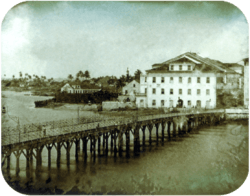 Photograph of a long wooden bridge crossing a river to a town with multi-storey white buildings and palm trees in the far background
