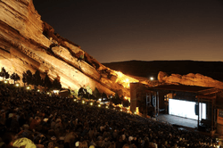 A shot of an outdoor amphitheater taken at dusk, looking down towards a brightly-lit stage. Large red cliffs are visible in the background, sloping down to the right. A large audience is present.