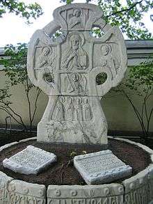 A large stone Russian cross with a figure carved on it, behind two stone tablets set into the ground