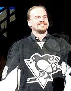 A Caucasian man smiling is shown from the waist up. He wears a black jersey with an anamorphic penguin playing ice hockey for the logo