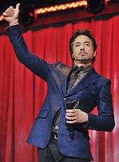 Robert Downey Jr with his right hand in the air giving a thumbs up while holding a microphone in his left hand