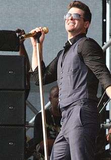 A picture of a brown-haired man from the shoulders up. He is wearing a black shirt and singing into a microphone.