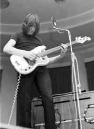 A monochrome image of Waters playing bass guitar. He has shoulder-length hair, black attire, and is standing in front of a microphone.