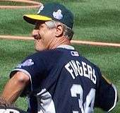 An older white male wearing a green cap with a yellow bill, a blue jersey, with the lettering "FINGERS" and a number 34 below it on the back, on a grass field.