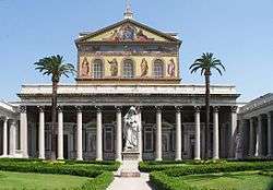 A courtyard with palm trees and a greater-than-lifesized statue of St. Paul holding a sword in front of the colossal portico of the basilica and a large mural covering the upper facade